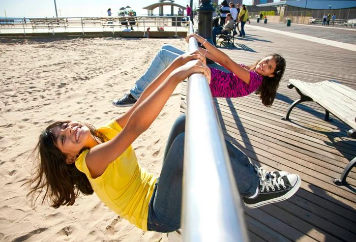 Two cheerful girls swing from a bar over the beach at Coney Island in New York City