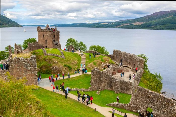 A bird's-eye view of a crumbling castle with numerous visitors by a loch