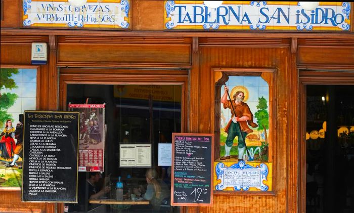 A front view of Taberna de San Isidro, a charming traditional tavern adorned with tiles, serving classic dishes, featuring chalkboard menus outside, and patrons dining at the front window