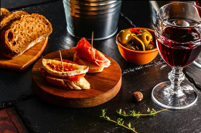 A close-up of a table displaying a plate of tapas (two slices of bread, one with ham and cheese, the other with just ham, secured with toothpicks), slices of plain bread, a bowl of olives, sprigs of thyme, and a glass of wine