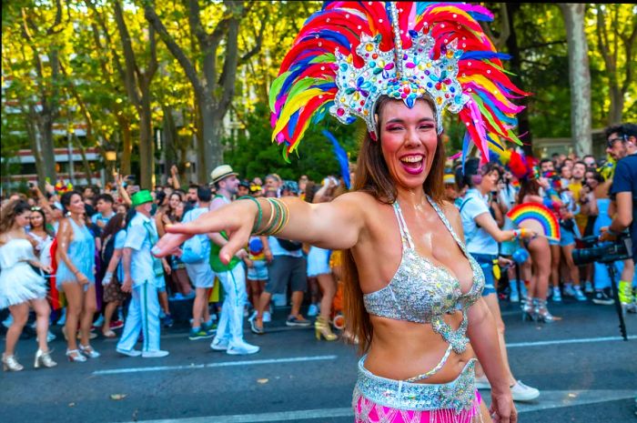 A parade dancer sporting a rainbow headdress smiles and poses for the camera. Title: Pride, Madrid