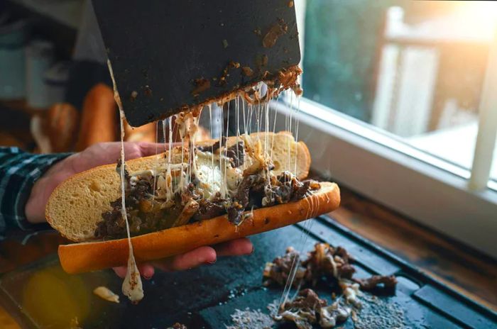 Hot, gooey, stringy cheese being pulled away from a Philly cheesesteak sandwich in a kitchen.