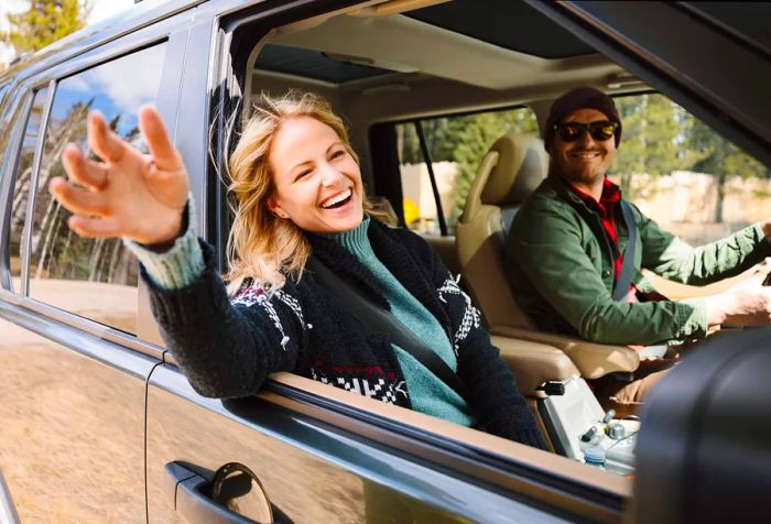 A couple inside a car, with the woman waving out the window while the man drives.