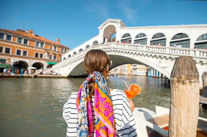 A young woman savoring a summer cocktail in front of the iconic Rialto Bridge in Venice, embodying the joy of a vacation in Italy with an Italian Aperol spritz in hand.