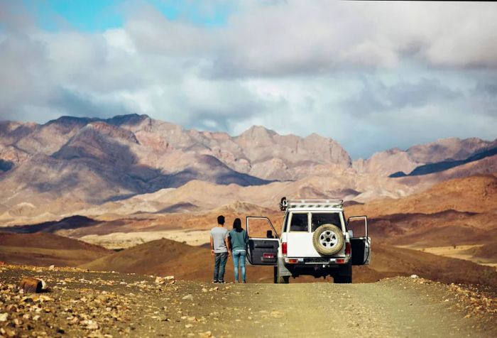 A couple admires the view from their vehicle, gazing at the rolling rocky mountains beneath a cloudy sky.