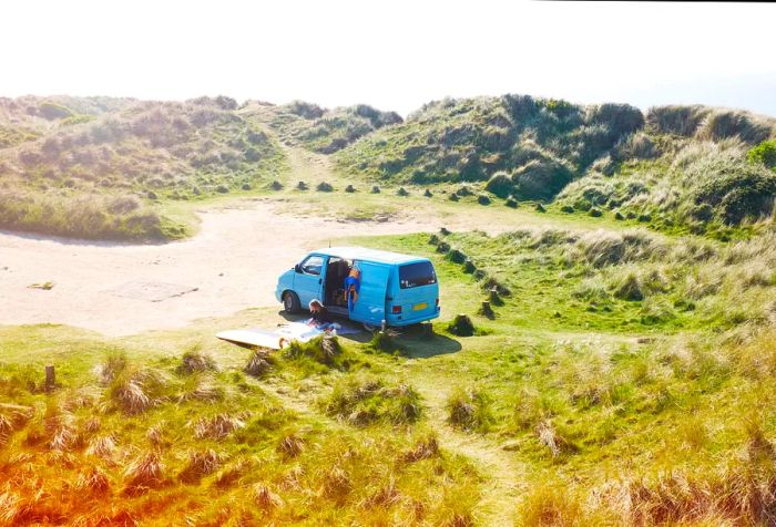 A person relaxes on a picnic mat next to a parked van amid grassy sand dunes.