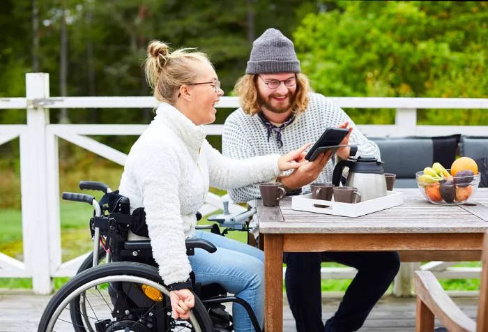 A joyful woman wearing a knitted white long-sleeve top sits in a wheelchair, pointing at a mobile device being shown to her by a man beside her.