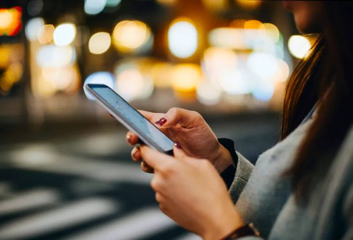 A woman is using her smartphone on a bustling sidewalk, with the vibrant lights of the city blurred in the background.