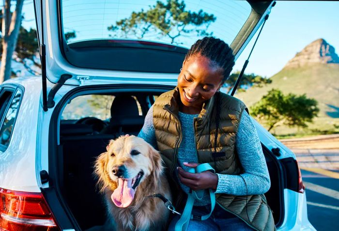 A cheerful woman perches on the trunk of her car, affectionately petting her dog.