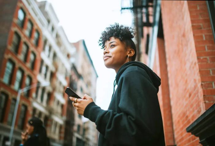 A woman with curly hair wearing a black hoodie checks her phone while standing on a sidewalk.