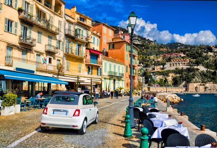 A street adorned with dining tables and outdoor cafés, bordered by buildings that overlook a body of water, all supported by a slope covered in structures.