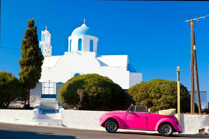 A pink Volkswagen Beetle parked in front of a chapel on Santorini, Greece