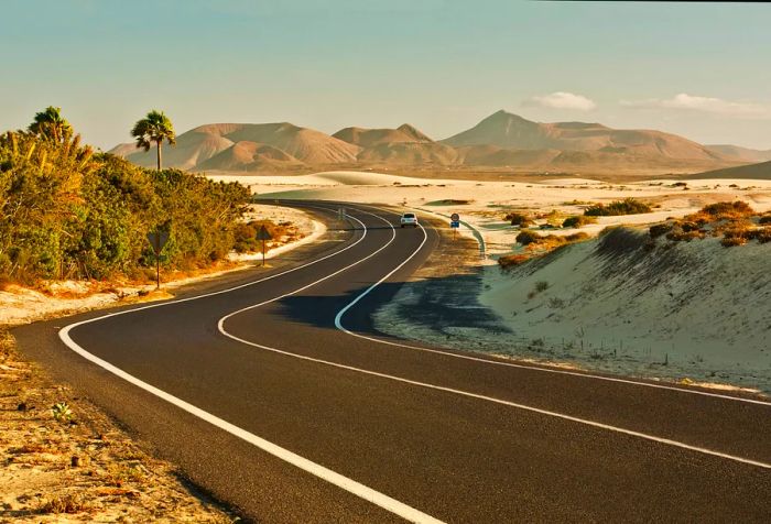 A car winds along a road flanked by grassy sand dunes, with mountains visible in the distance.