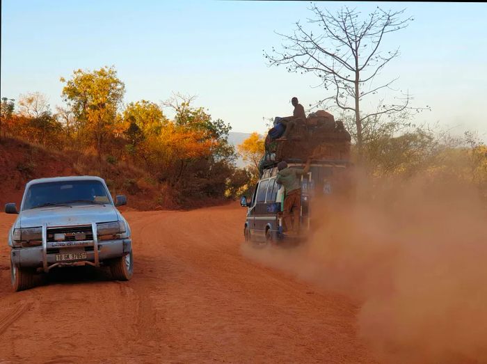 A car is parked beside a dusty red road in West Africa.