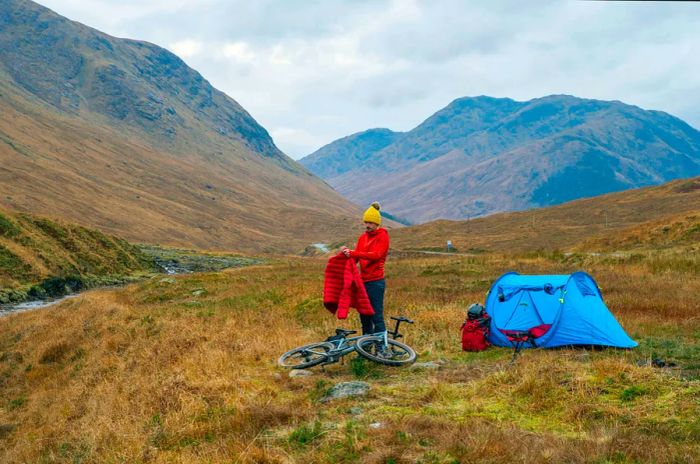 A solo camper stands between his bike and his tent amidst the stunning scenery of the Scottish Highlands