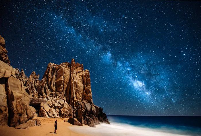 A person gazes up at the stars in a serene sky from a picturesque beach framed by steep, rugged cliffs.