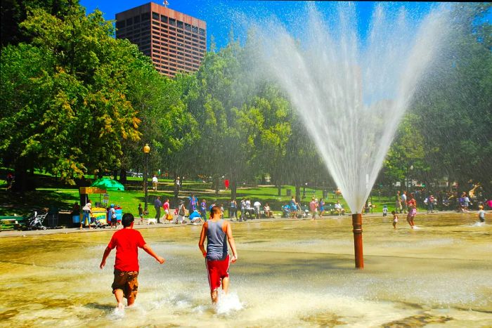 Two teenagers play in a shallow outdoor pool, enjoying a large jet of water shooting high into the sky on a sunny day.