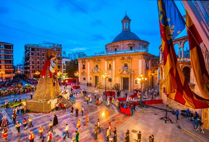 A vibrant festival unfolds in a plaza, featuring a massive wooden statue of the Virgin Mary adorned with a red mantle. People gather around, offering flowers, with many dressed in traditional costumes.