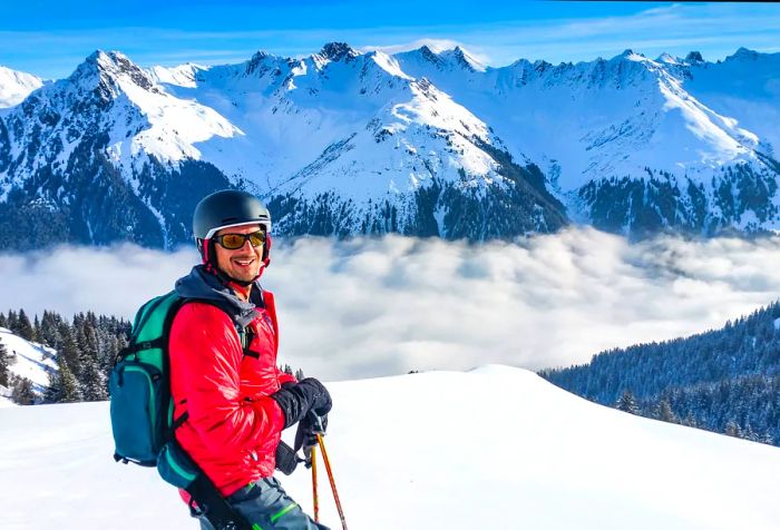 A cheerful skier clad in a red jacket and helmet stands atop a snowy hill, surrounded by dense fog at the base of snow-covered mountains.