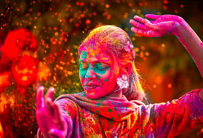 A woman adorned with large earrings dances joyfully, splashed with bright dye powder during the Holi festival.