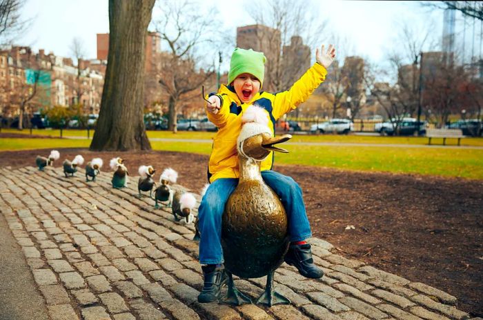 A small child beams with joy while sitting atop a bronze duck sculpture.