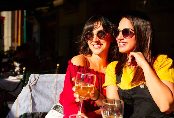 Two joyful women relax on a bench, enjoying drinks and soaking up the sunshine.