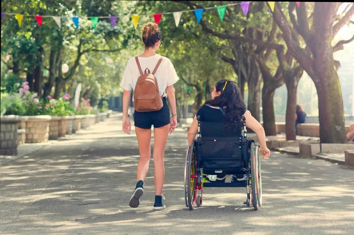 A young woman strolls beside her friend in a wheelchair in the Italian town of Tempio Pausania, where the walkway is wide and lined with greenery.