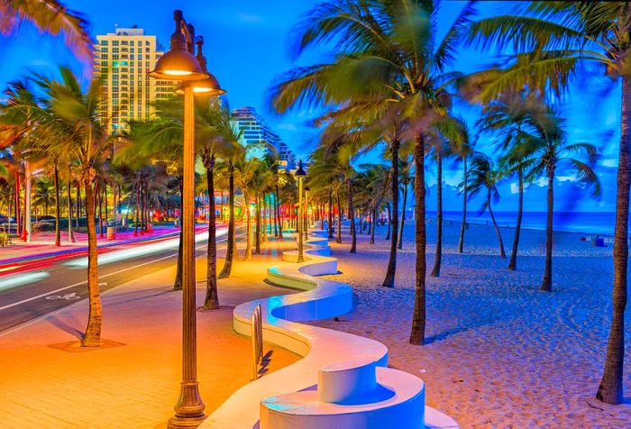 A long, curving white concrete bench nestled among palm trees and lampposts that line the seaside road against a backdrop of clear blue skies.