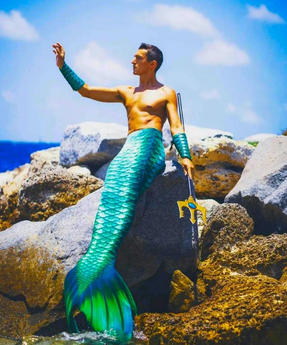 Merman Andrew perches on a rock, his right arm raised, wielding a three-pronged trident, with his green mermaid tail cascading down towards the beach.