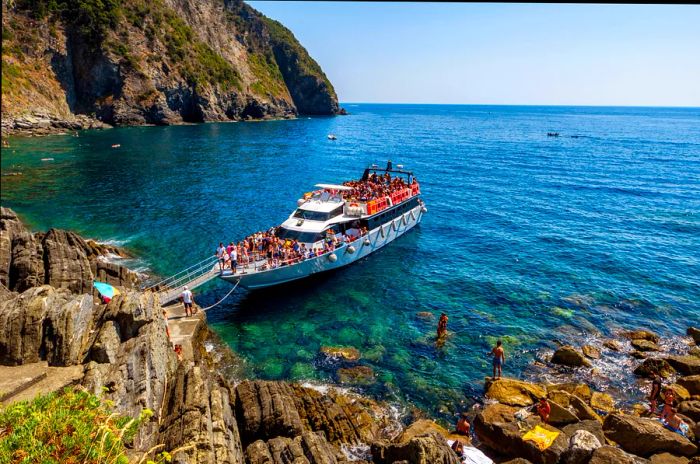 A ferry boat drifts in the clear blue waters off the coast of Riomaggiore in Cinque Terre, bustling with passengers on deck.