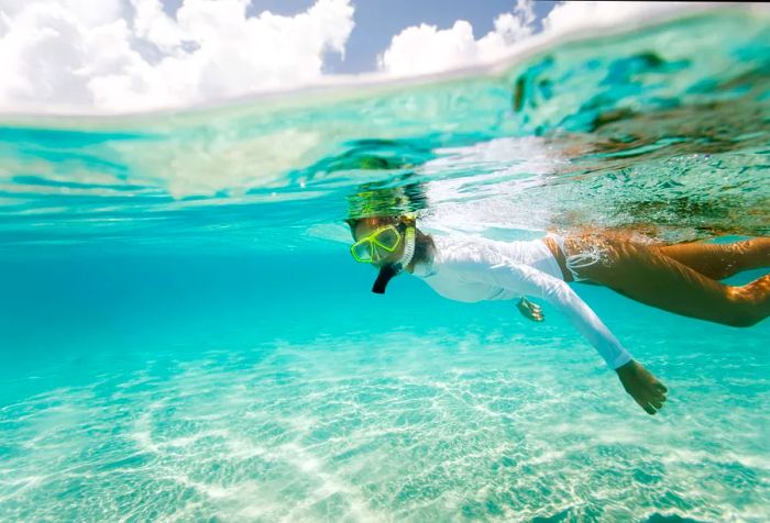 A woman wearing a white rash guard snorkels in crystal-clear waters.