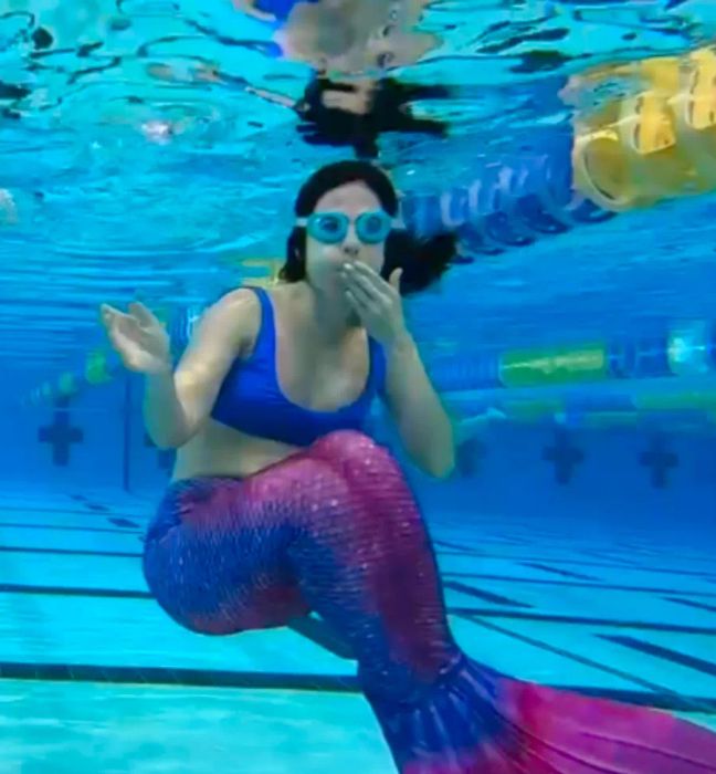 The writer, fully equipped in mermaiding gear, is submerged underwater in a swimming pool with goggles on, trying out the hobby for the first time.