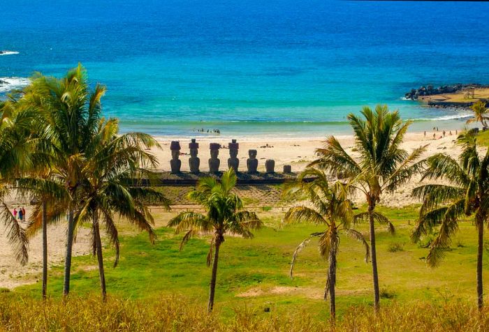 Coconut palms dot a grassy area along the beach, accompanied by a row of intricately carved figures in the sand.