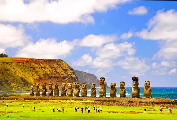 The famous 15 Moai statues, massive stone figures crafted by the Rapa Nui people, proudly line the coast, with visitors wandering the surrounding terrain and a stunning coastal cliff as their backdrop.