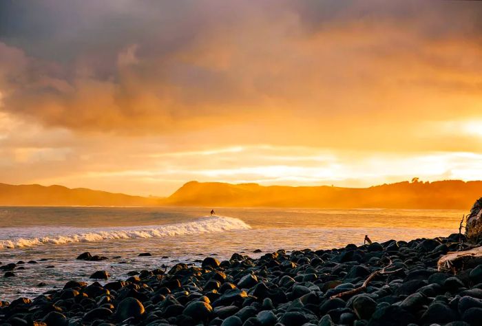 A scenic view of a rocky beach at sunset, showcasing waves and surfers. Raglan, New Zealand