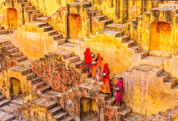 Indian women elegantly carry water from a stepwell, skillfully navigating the series of stairs, while the weathered paint of the surroundings adds a rustic allure.