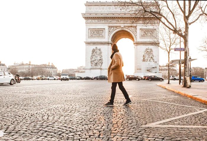 A man dressed in a winter coat strolls along the cobblestone streets in front of the Arc de Triomphe.