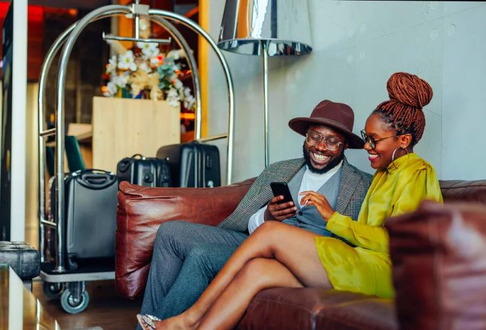 A well-off couple relaxes in the hotel while waiting to be escorted to their luxurious suite. They sit on a sofa with their luggage on a cart, both using a smartphone.