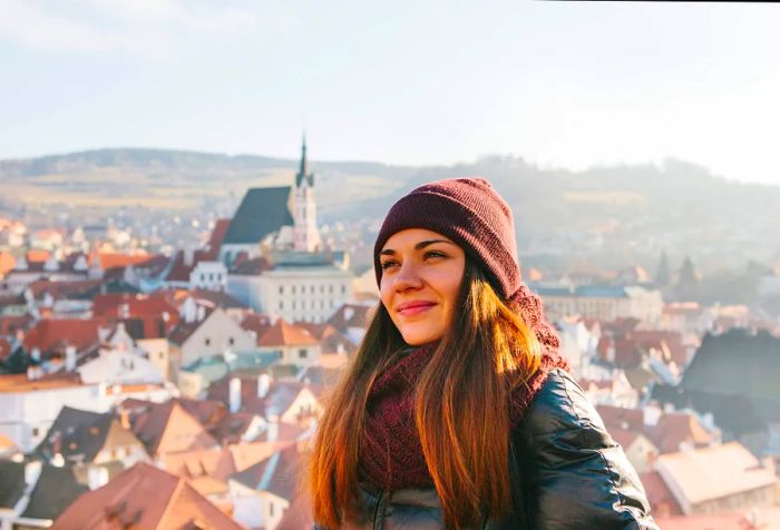 A cheerful brunette girl in a winter hat and cozy attire with a charming town in the background.