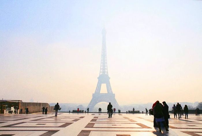 A plaza with a patterned tile design, where a large crowd gathers to admire the view of the Eiffel Tower shrouded in dense mist.