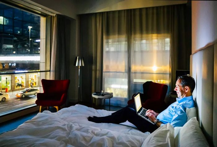 A businessman is seated on a hotel bed, working diligently on his laptop.