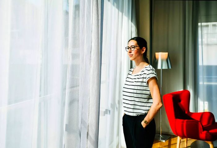 A woman wearing a black and white striped shirt stands by the window, hands tucked into her pant pockets.