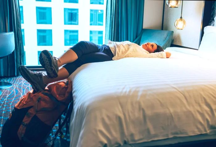 A female backpacker relaxes comfortably on the white bed in her hotel room.