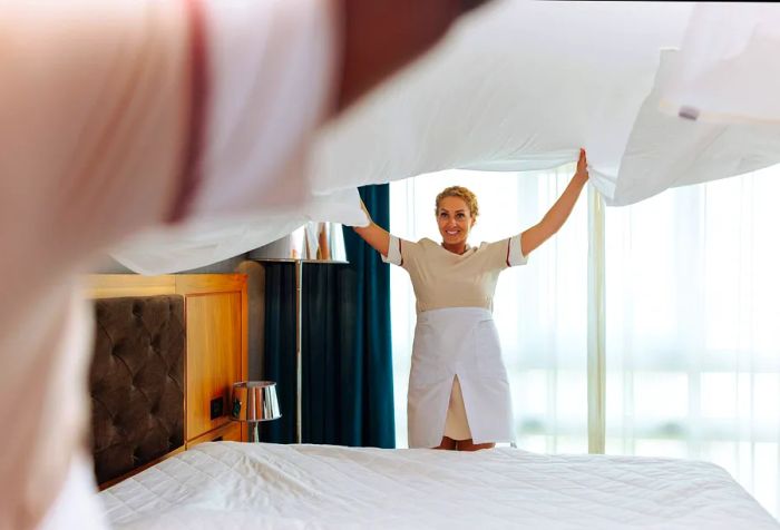 Two young multiracial hotel attendants are joyfully lifting a clean bed sheet into the air together.