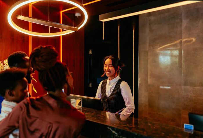 A young Asian receptionist is engaging with her African American guests at the front desk of an upscale hotel.