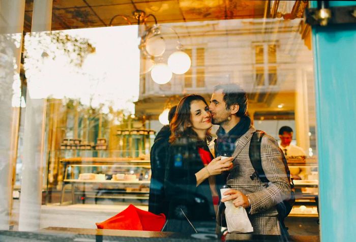 A person sharing a kiss on another's cheek through a glass partition at a café.