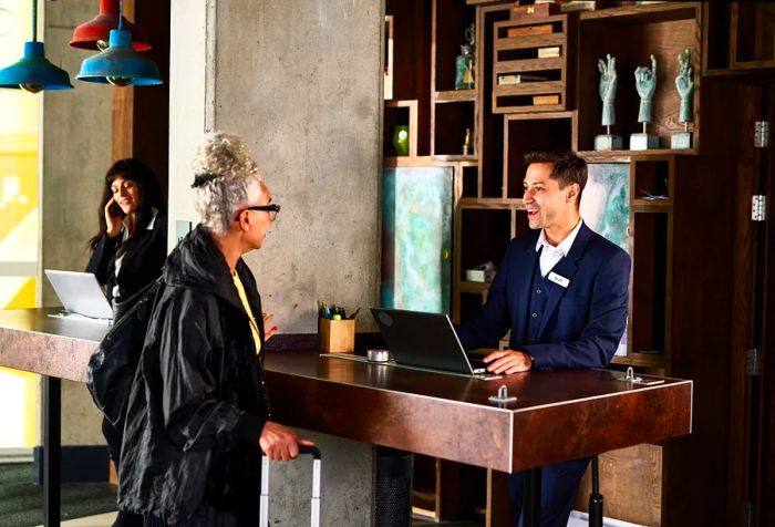 A male hotel receptionist dressed in a navy suit speaks with an elderly woman at the front desk.