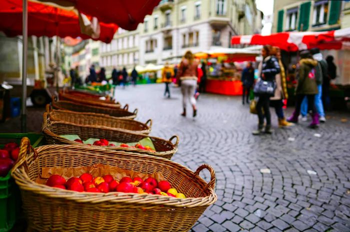 A fruit and vegetable stall in a cobblestone square bustling with activity on market day as people stroll by.