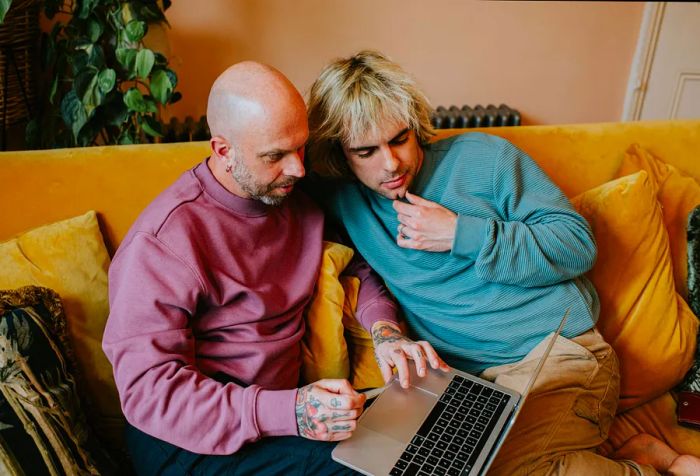 A male couple shares a laptop while relaxing on a yellow sofa.