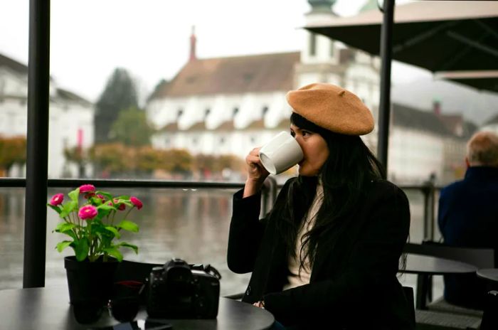 A woman sits at an outdoor table in Lausanne, sipping from a mug.